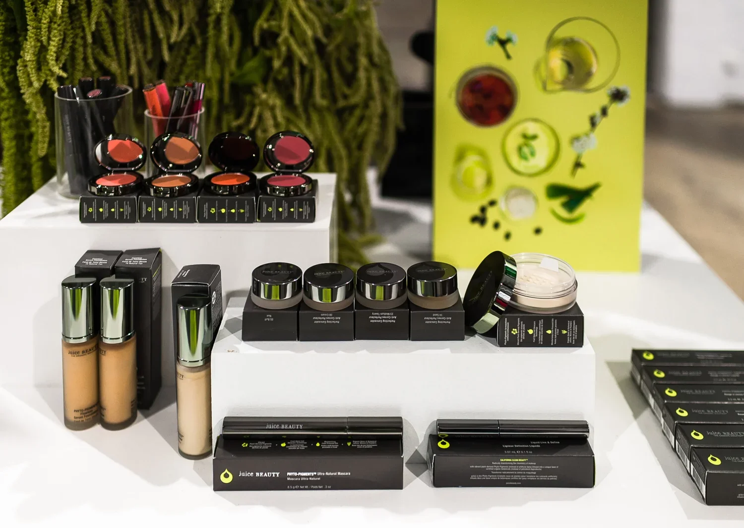 Display of Juice Beauty skincare and makeup products, including foundations, mascaras, and lip products, on a white table with a green decorative backdrop.