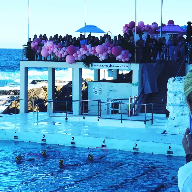 People gathered on a rooftop terrace decorated with pink and purple balloons, overlooking a pool with flower-shaped floating devices, near the ocean.
