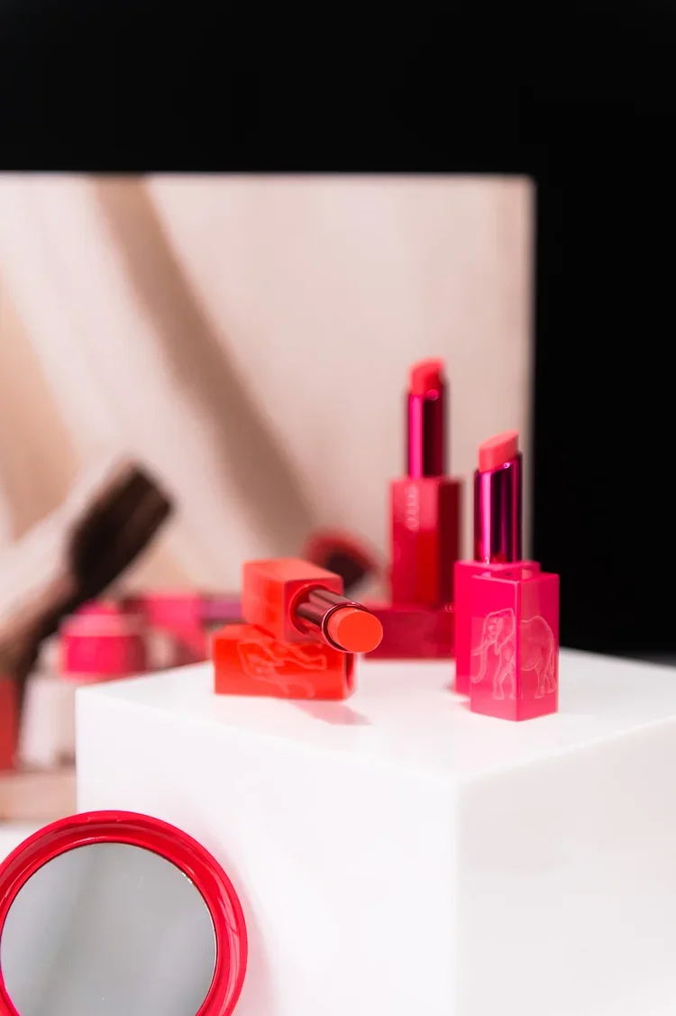 Close-up of three pink and orange lipsticks displayed on a white pedestal with a mirror in the background.