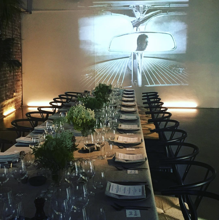 A long dining table set for a formal event, with black chairs, glassware, napkins, and floral centerpieces. A black and white image of a person driving a vintage car is projected on the wall at the end of the room.