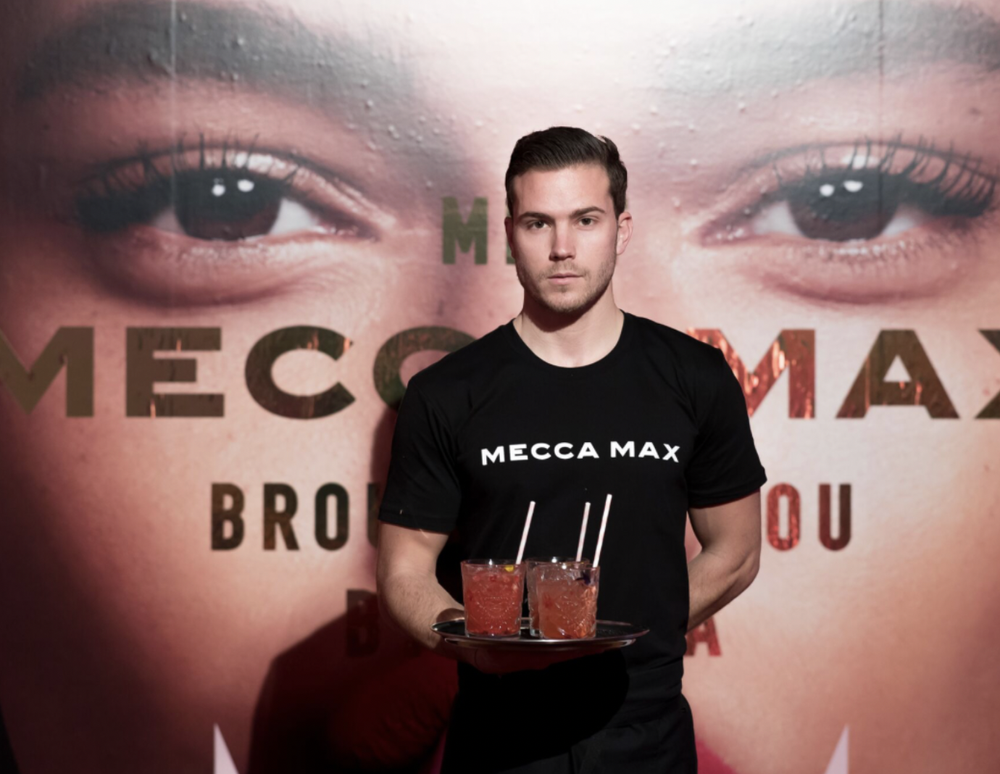 A man in a black t-shirt with "Mecca Max" written on it is holding a tray with two glasses of red drink with straws, standing in front of a large promotional poster featuring a close-up of a woman's face with bold makeup.