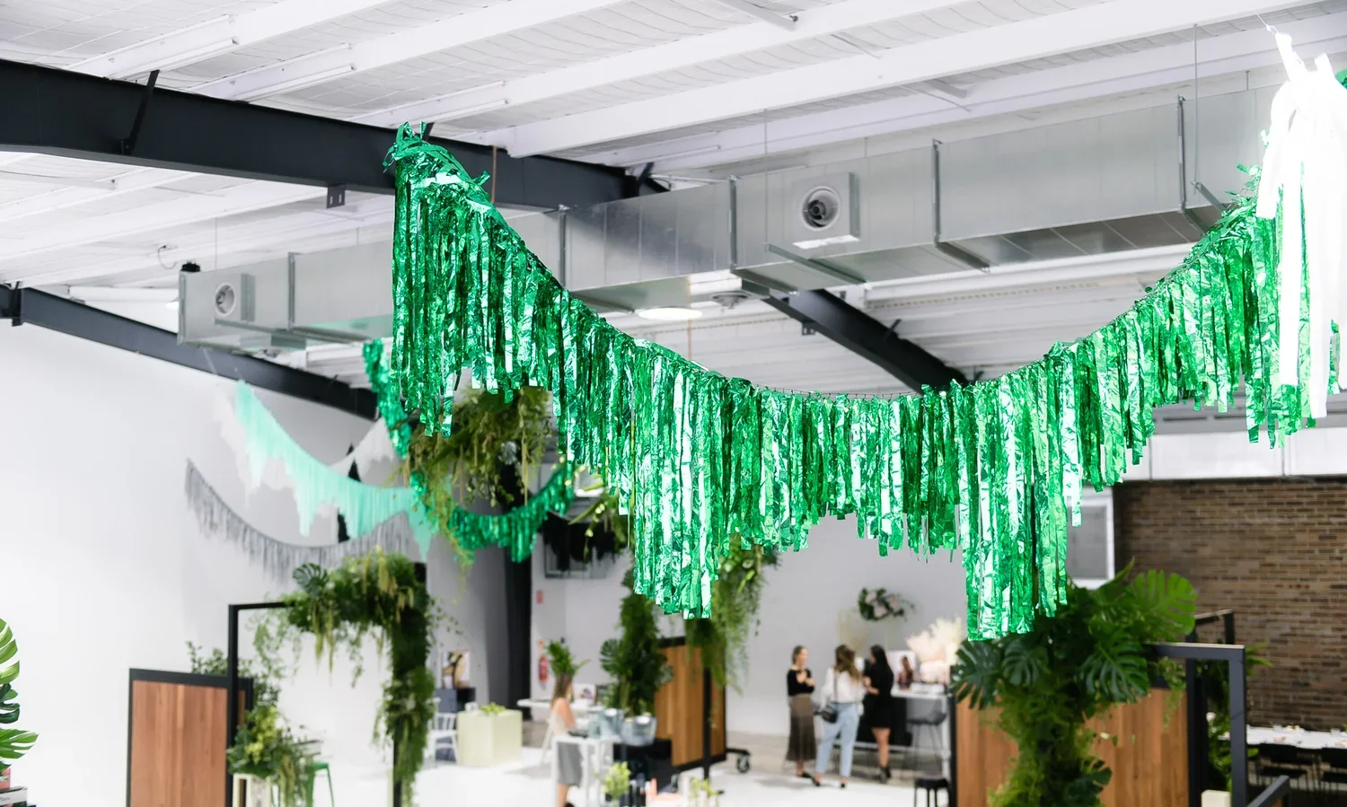 Indoor space decorated with green tissue paper banners hanging from the ceiling, with people gathered around tables and a counter in the background, surrounded by plants.
