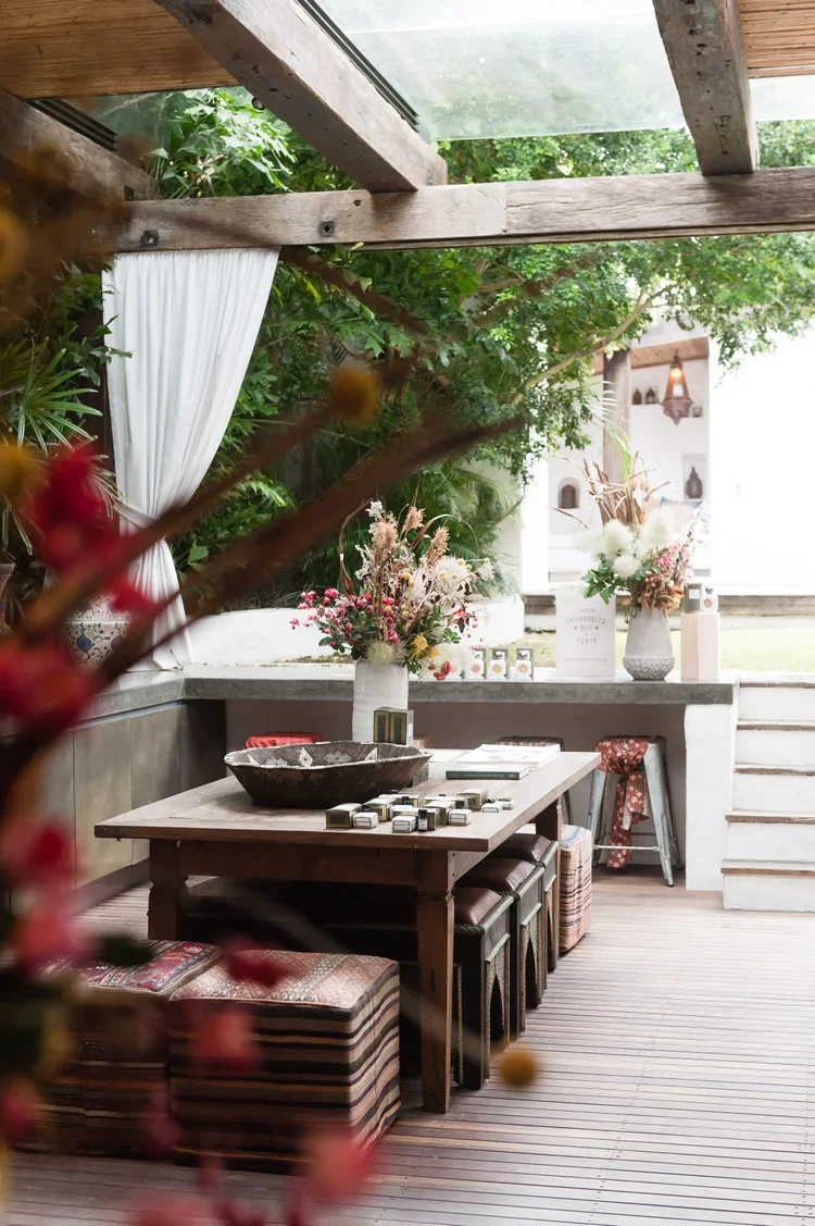 Indoor patio with wooden table, floral arrangements, patterned stools, and lush green plants in the background.