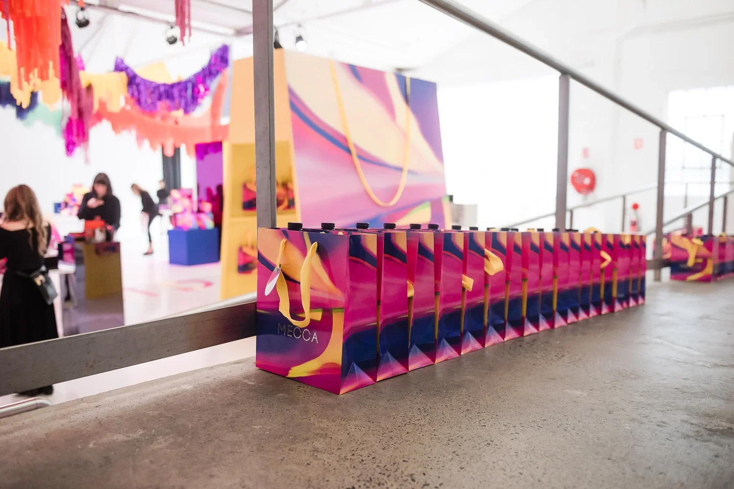 Series of colorful shopping bags with yellow handles, lined up on a concrete ledge at an event space, with a vibrant pink, purple, and yellow abstract mural in the background.
