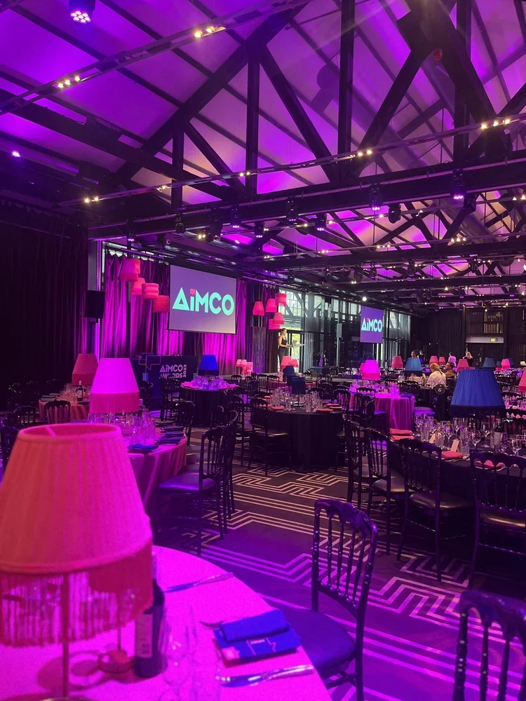 Event hall decorated with pink and purple lighting, multiple tables with chairs, table settings with glasses and utensils, large screens displaying 'AIMCO', and a stage with a performer.