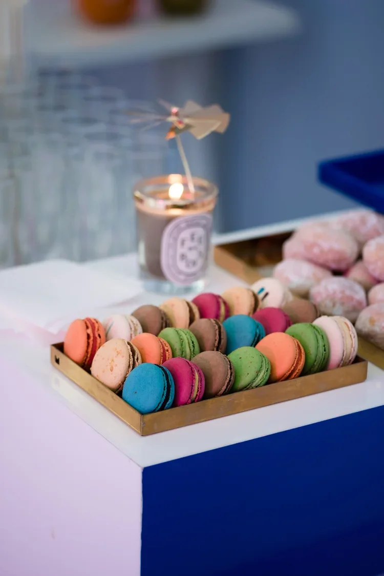 Colorful macarons arranged in a tray on a white table with a candle and pink candies in the background.
