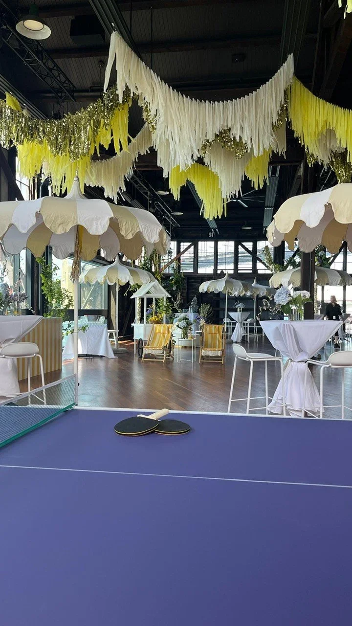 Indoor event space decorated with yellow and white hanging paper tassels, white parasols, tables with white tablecloths and floral centerpieces, and a table tennis paddle set in the foreground.
