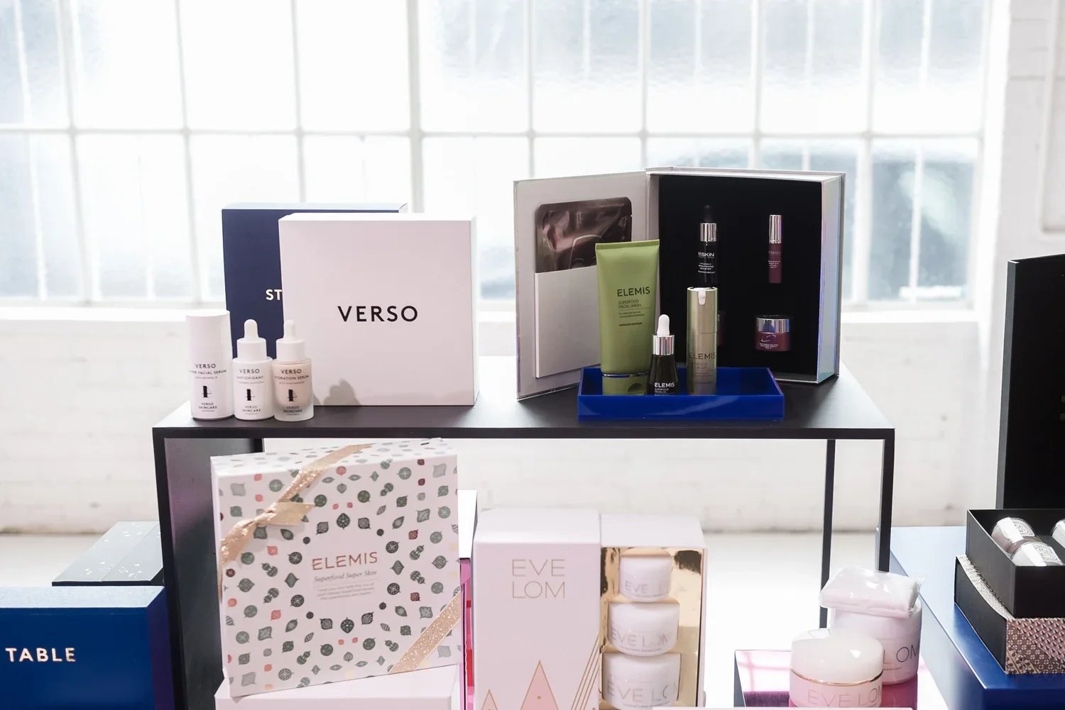Display of skincare and beauty products on tables, including boxed sets, bottles, and jars from brands like Verso, Elemis, and Eve Lom, in a bright, modern room with large windows.