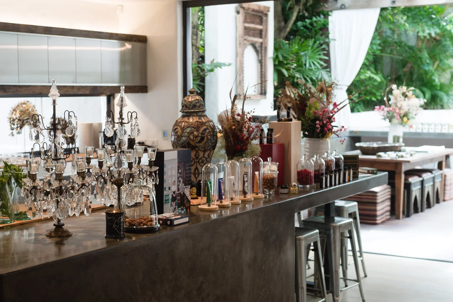 Decorated bar with crystal chandelier, vials of spices or herbs, flowers in vases, and natural light from large open windows with outdoor greenery.