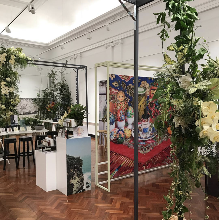 An art exhibit with floral arrangements, colorful painted eggs on a tapestry, and various artistic displays in a gallery with wooden floors and white walls.