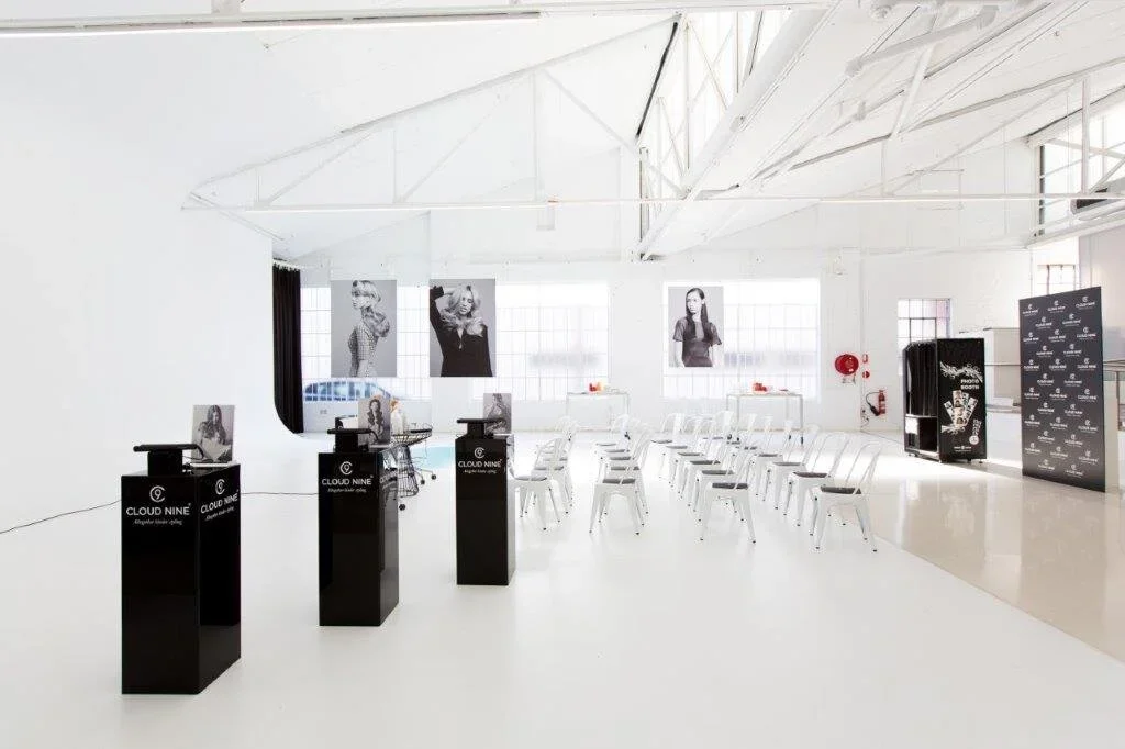 White event space with chairs arranged in rows, black presentation stands, large black and white photographs of women hanging on the wall, and a black vending machine, with a bright windowed background.