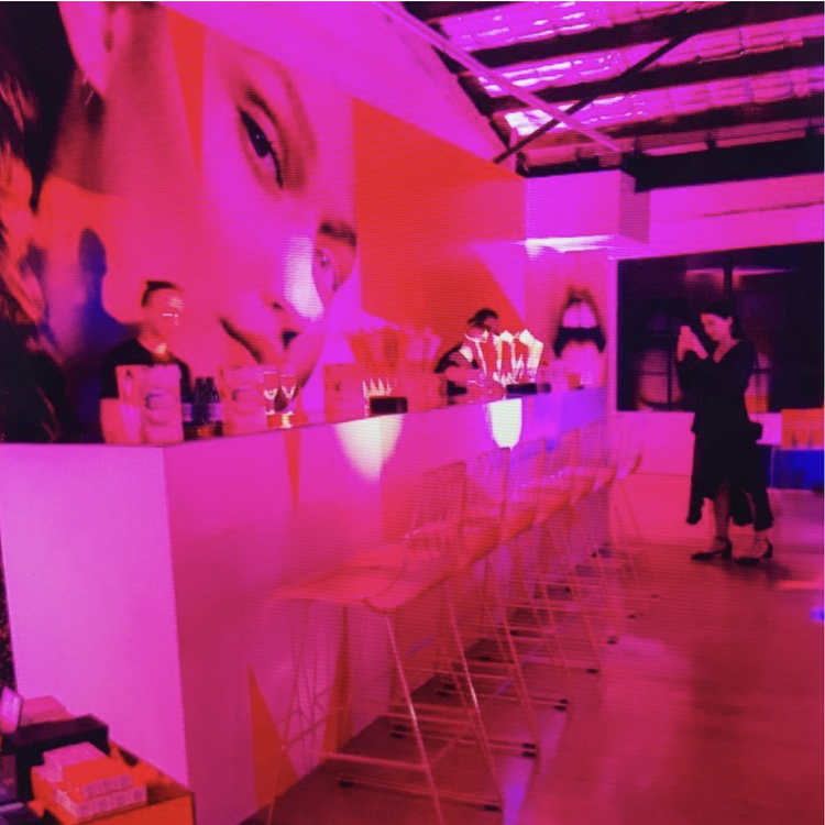 A modern bar with a large pink and purple poster of a woman's face on the wall. There are empty white bar stools lined up along the counter. A person in black is taking a photo or video of two other people at the bar, with drinks and decorations on t