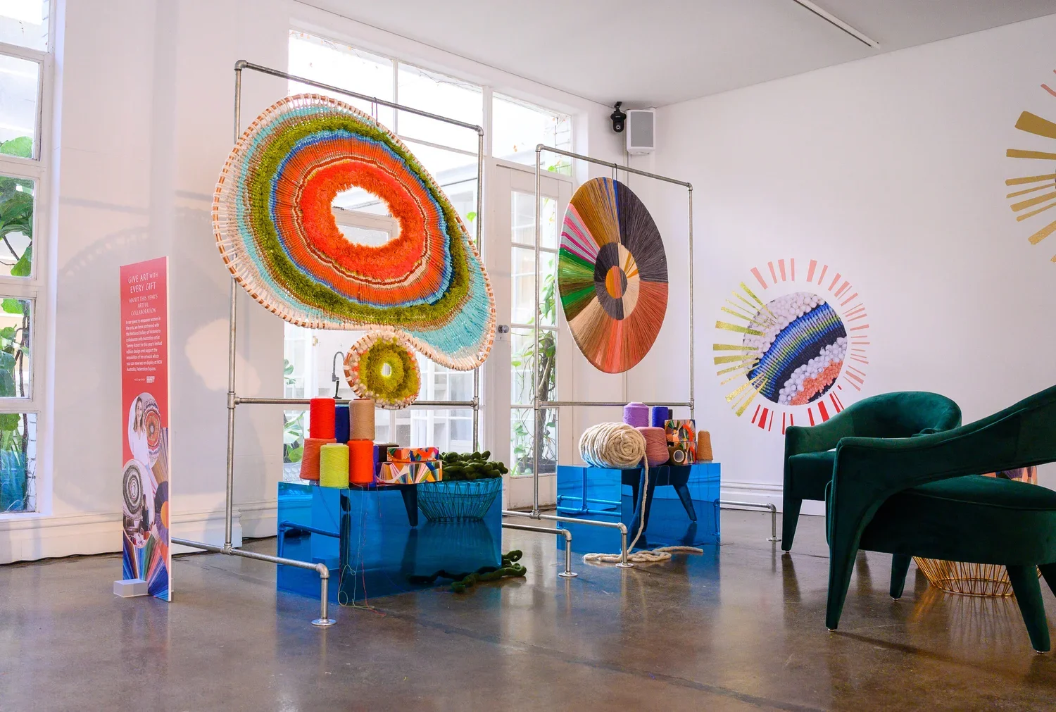 Indoor art installation with colorful woven circular pieces displayed on metal frames, with yarn and fiber art supplies on blue tables, and green velvet chairs nearby.