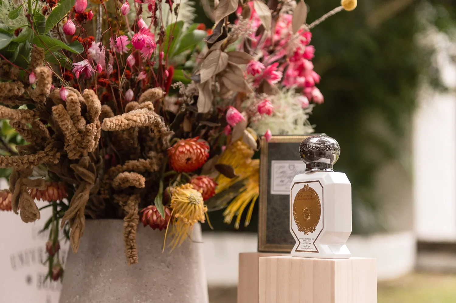 A floral arrangement with pink, yellow, and brown dried flowers in a concrete vase, placed next to a white perfume bottle with a silver cap on a wooden pedestal, with a box behind it.