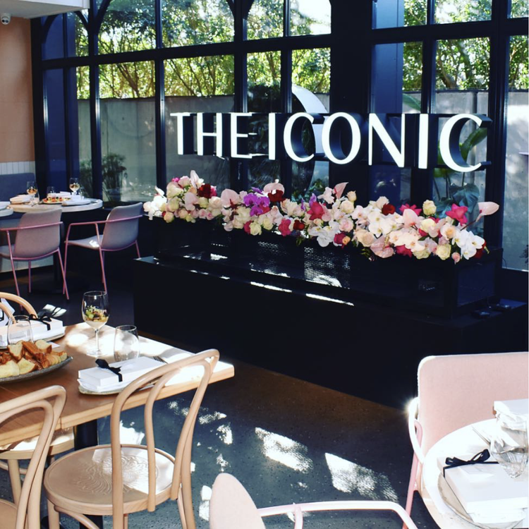 Dining area with round tables, chairs, and a large window, decorated with a sign reading 'The Iconic' and a floral arrangement.