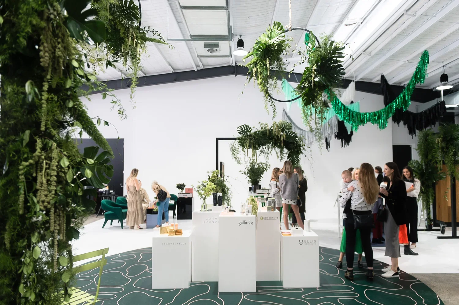 An indoor event space decorated with greenery and hanging green and black fringed garlands. Several people are gathered around booths and displays, with some wearing casual attire and others in business casual clothing. The space features a white wal