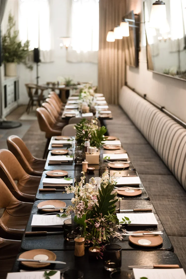 A long dining table set for a meal with white flowers and greenery in the center, placed in a well-lit room with white and brown curtains, leather chairs, and place settings including plates, utensils, and glasses.