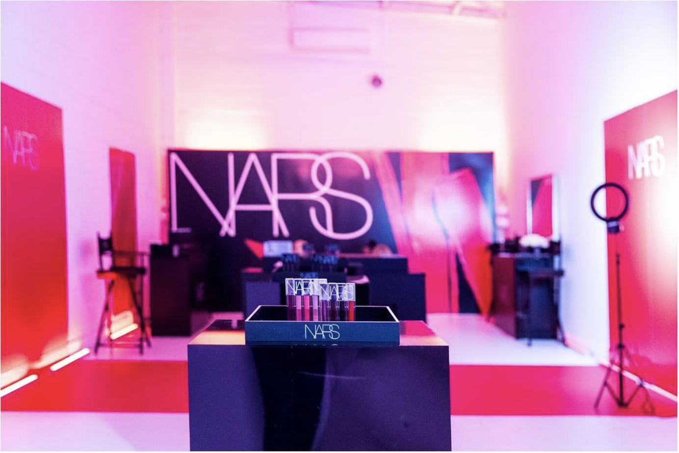 NARS beauty product display at an event with pink and purple lighting, featuring the NARS logo on a large backdrop and a ring light on a stand to the right.