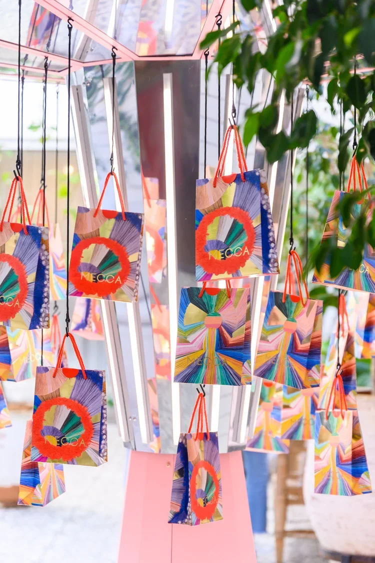 Colorful paper gift bags hanging from hooks on a metal rack, with some green leaves partially obscuring the view.