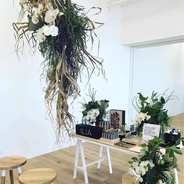 Display table with Ilia skincare products, surrounded by white flowers and greenery, with a hanging floral arrangement above on a wooden floor and plain white wall background.