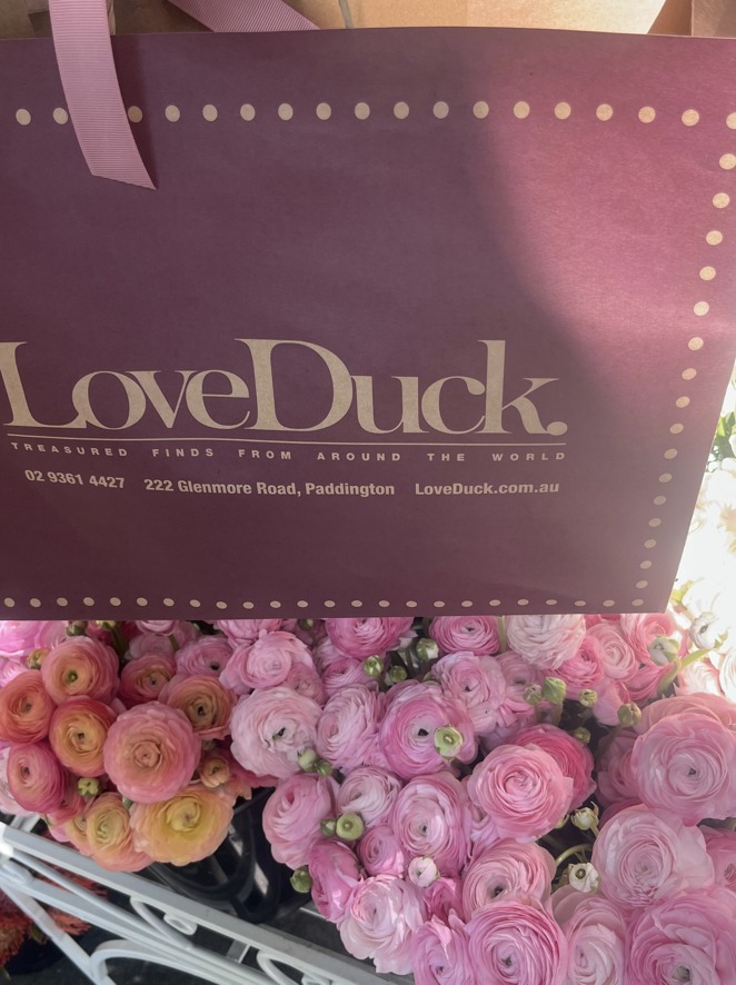 A pink Love Duck shopping bag hanging above a display of pink ranunculus flowers.