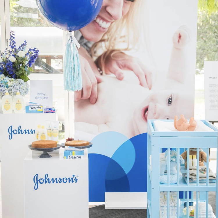 Display at a baby product event with Johnson's branding, baby skincare products, a cake, and a crib with baby supplies, with a large poster of a smiling woman and a baby in the background.