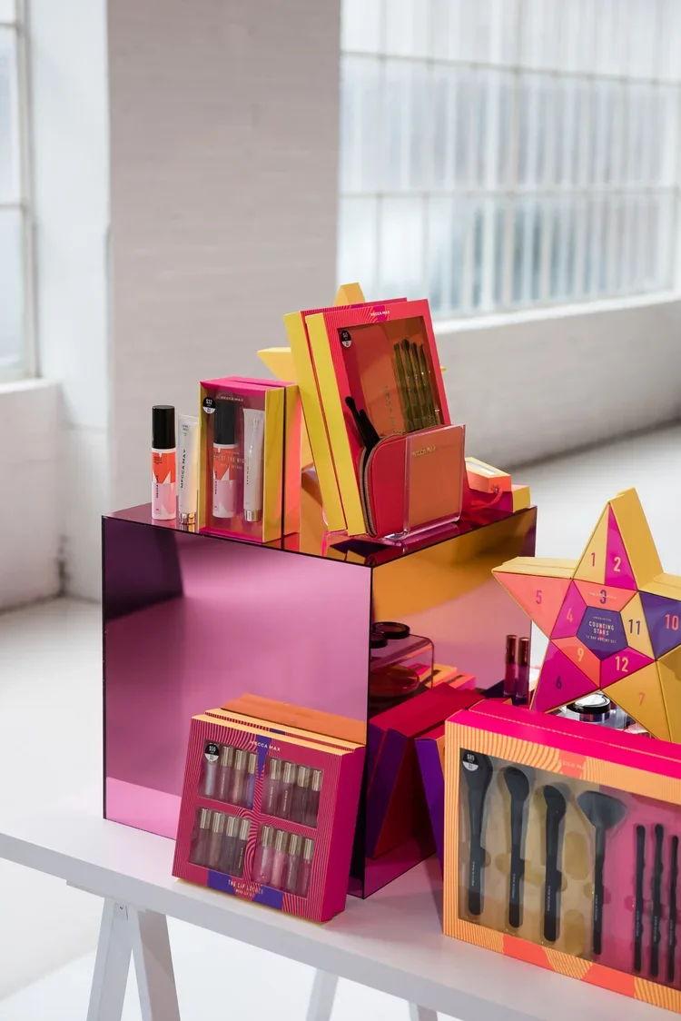 Colorful makeup and beauty products arranged on a pink reflective surface and a white table with a modern, bright background.