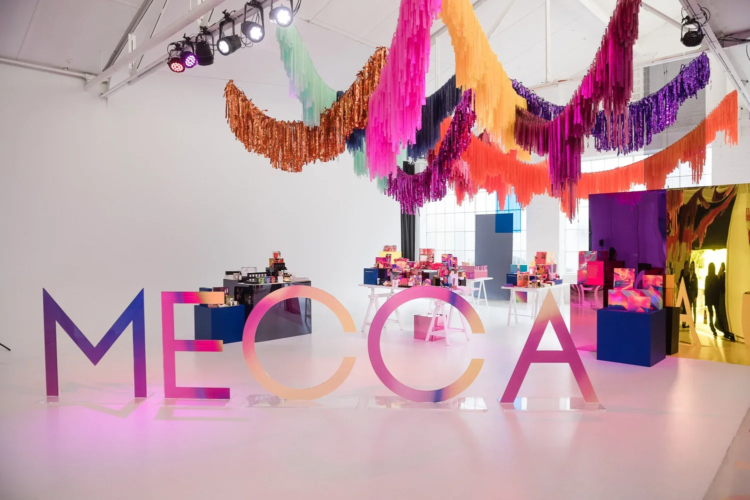 An indoor event space with large colorful 'MECCA' letters in the foreground, decorated with pink and purple flowers, and hanging multicolored paper streamers from the ceiling. Various tables display products or items, and people are seen in the backg