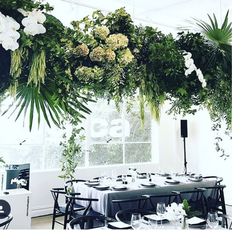 A dining table set for a formal event with black chairs, white tablecloth, glassware, and black napkins, surrounded by lush green floral and leafy decorations hanging from the ceiling, with large windows in the background.