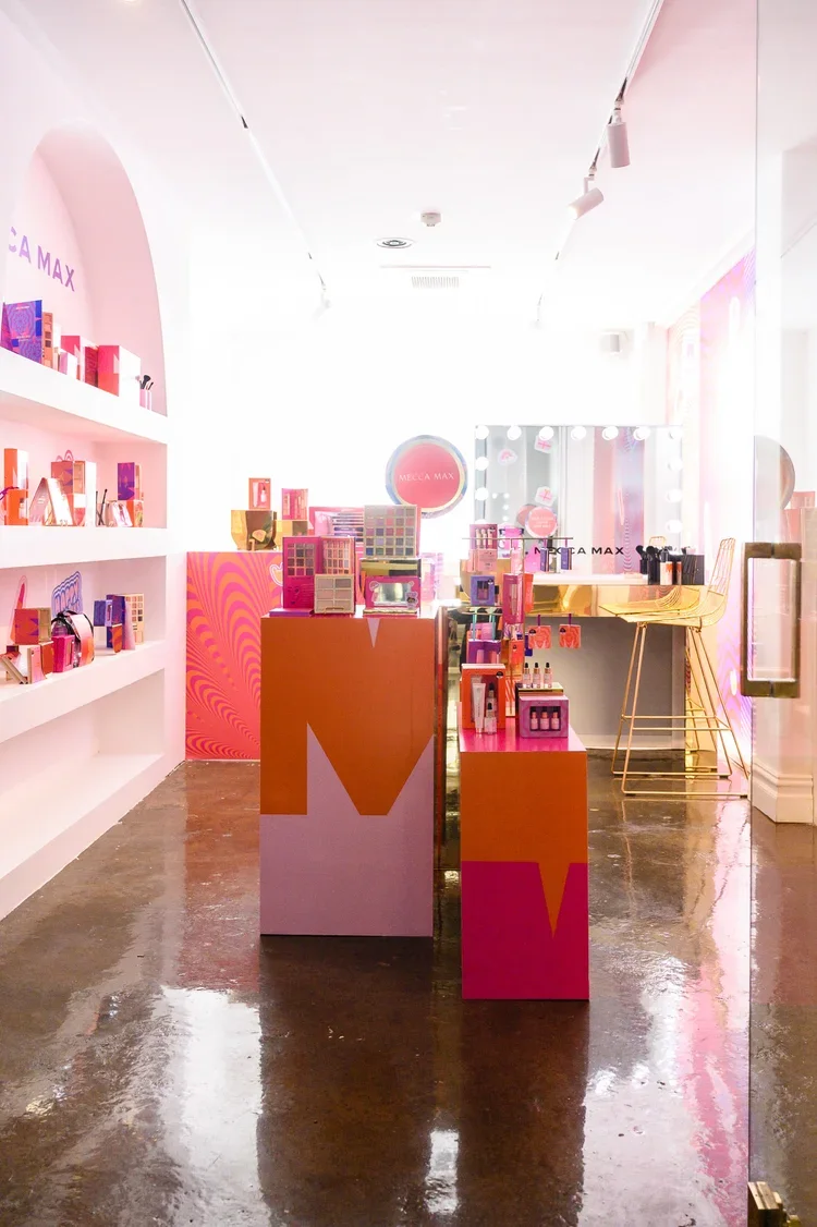 Inside a brightly lit cosmetics store with pink and orange displays, shelves with makeup products, and a large mirror with lights around it.