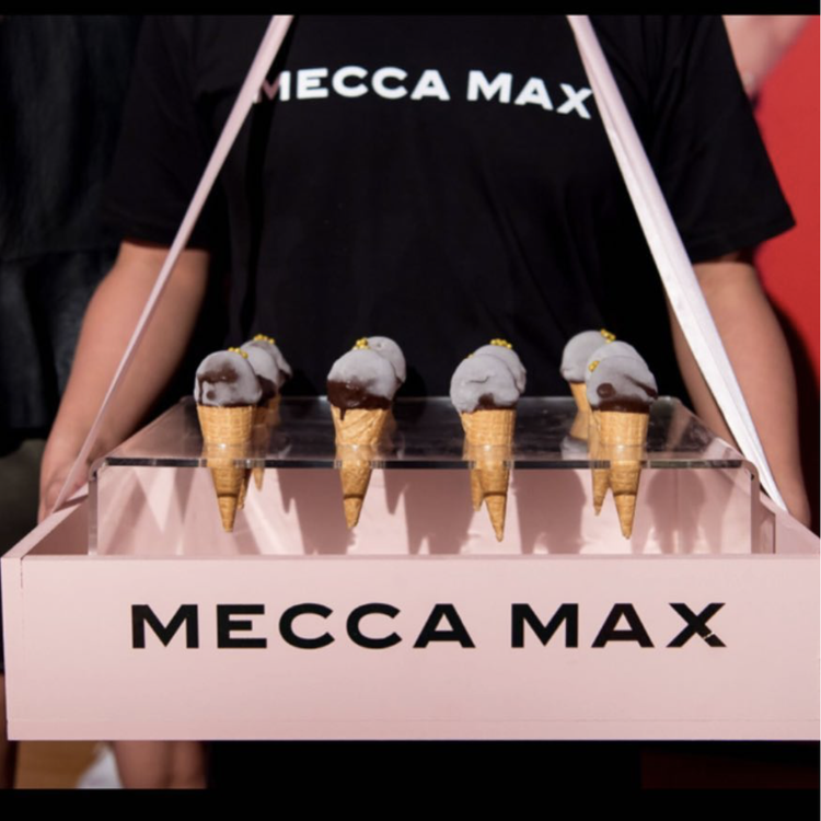 Person holding a pink display box with five ice cream cones topped with gray ice cream and small yellow decorations. The person is wearing a black shirt with the words 'MECCA MAX' printed on it.
