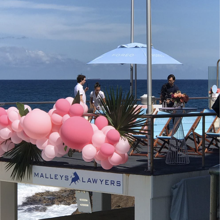 Beachside setting with pink balloons and greenery decorations, two women and one man standing, an umbrella, and a man preparing flowers on a deck overlooking the ocean.