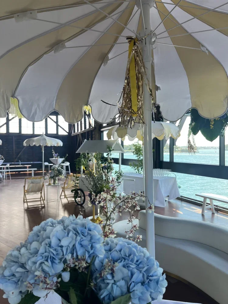 Decorative setup with white and yellow umbrellas, floral arrangements, and waterfront view inside a glass-walled venue.