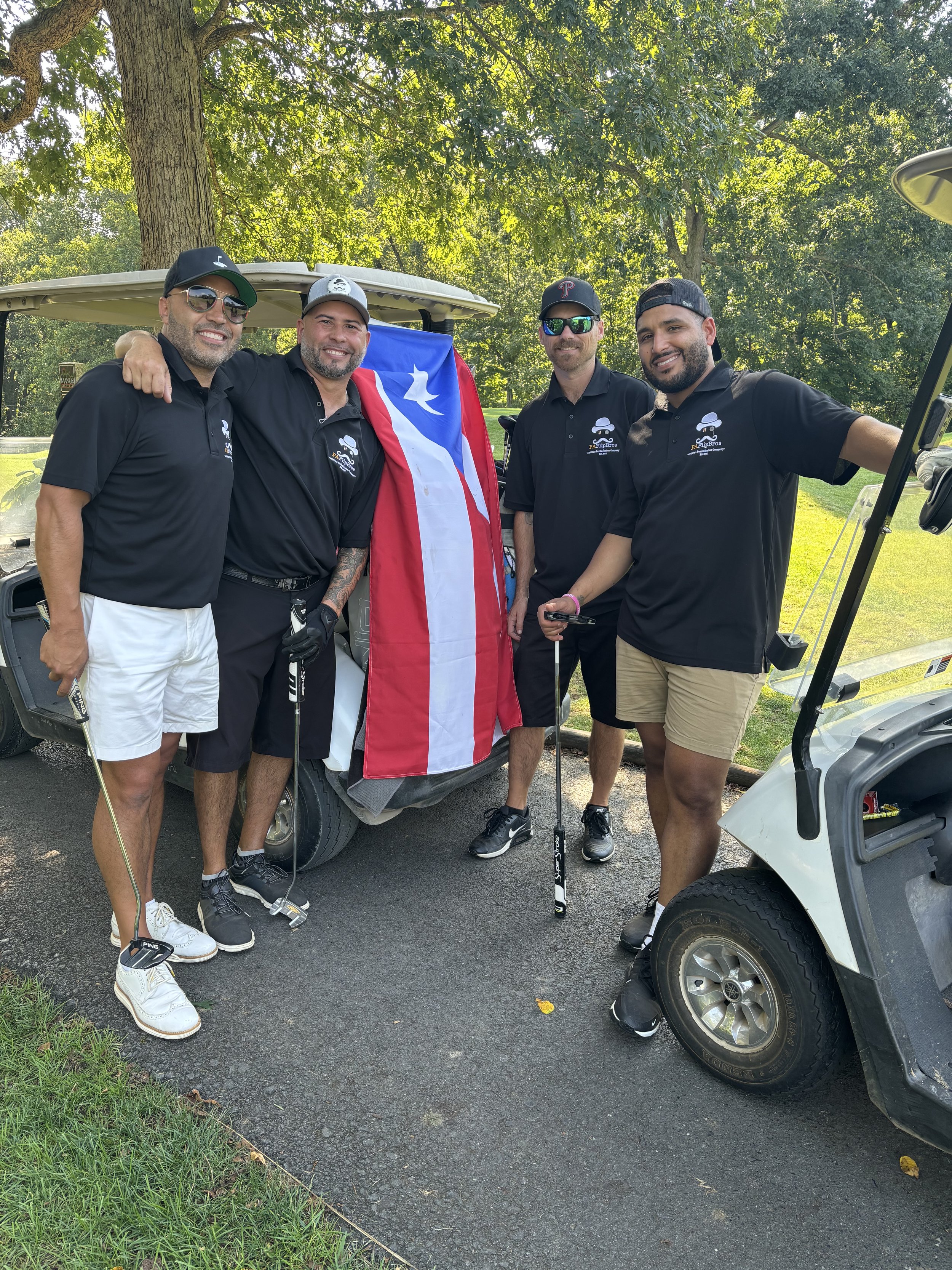 Four men wearing black polo shirts and shorts stand near golf carts on a golf course. They are holding golf clubs and a flag, likely Puerto Rican, draped over one of the golf carts. Trees and grass are visible in the background.