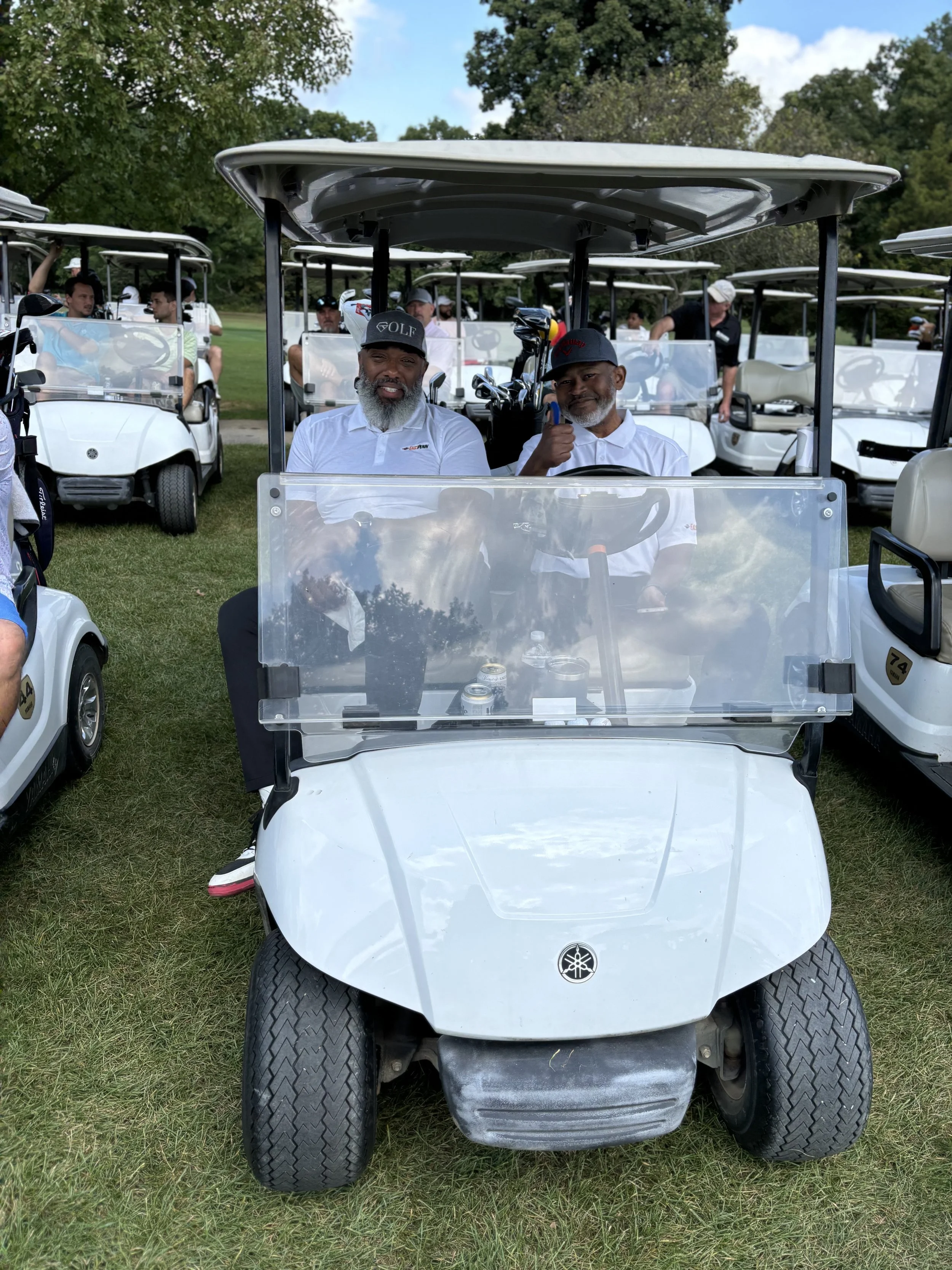 A group of people sitting in golf carts on a golf course. Two men are in the foreground cart, both wearing hats and white shirts, with one giving a thumbs-up. More people are visible in the background in other golf carts.