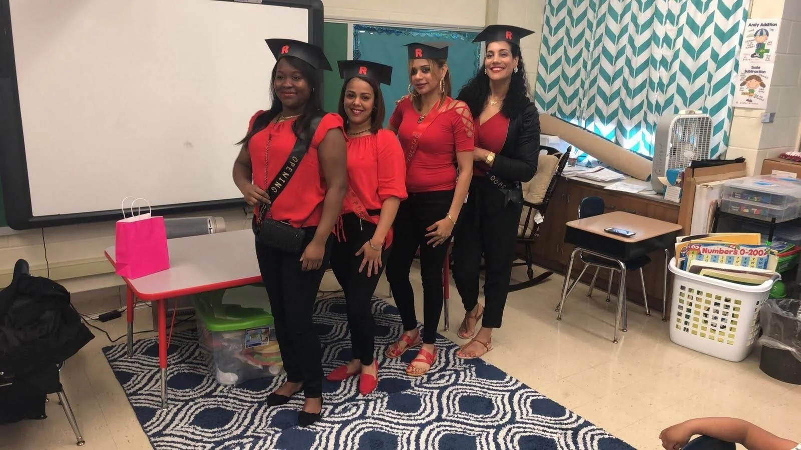 Group of four women in a classroom wearing graduation caps and red tops, posing in front of a whiteboard. A table with a pink gift bag is in the background.