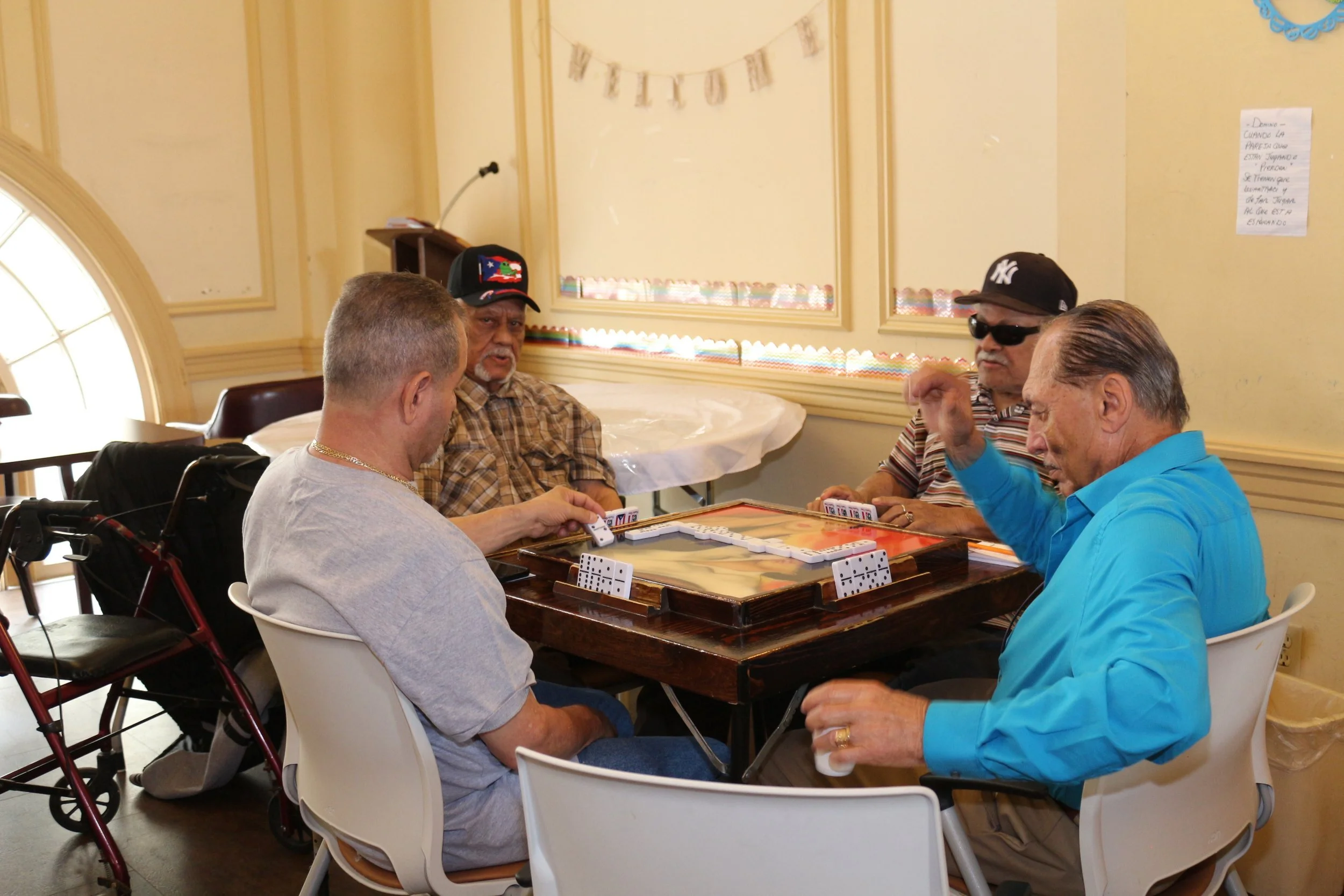 Group of elderly men playing dominoes around a table in a community room.
