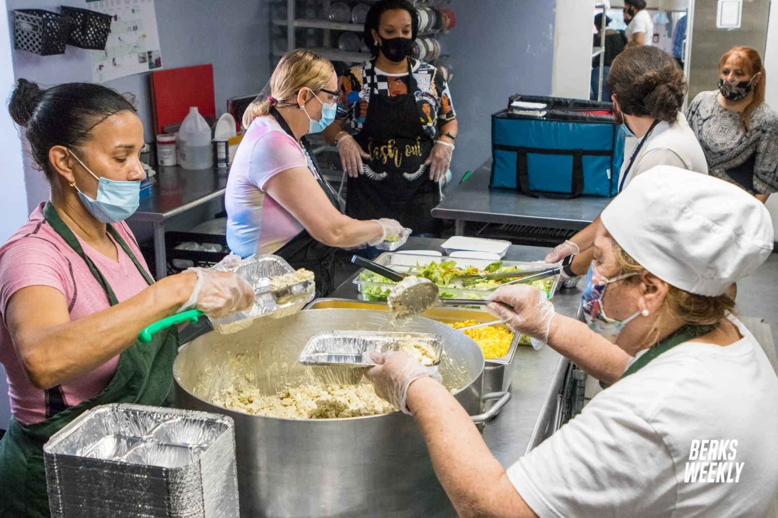 Volunteers preparing meals in a kitchen, wearing face masks and gloves, scooping food into containers.