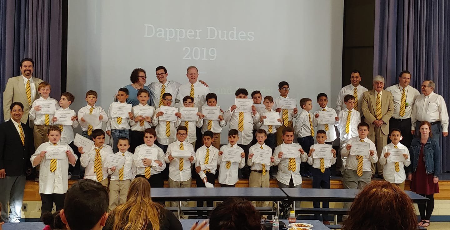 A group photo of boys and adults in white shirts with yellow striped ties, holding certificates in front of a screen displaying 'Dapper Dudes 2019.'