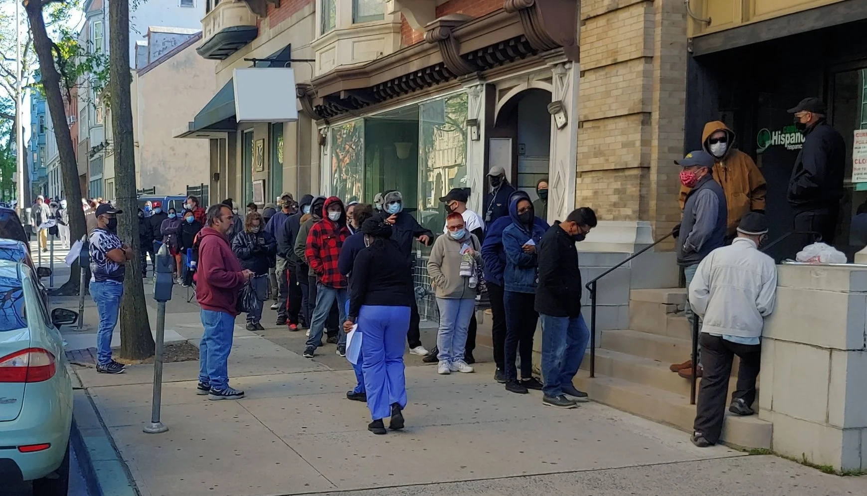 People in line on a city sidewalk wearing masks, outside a building with a Hispanic center sign.