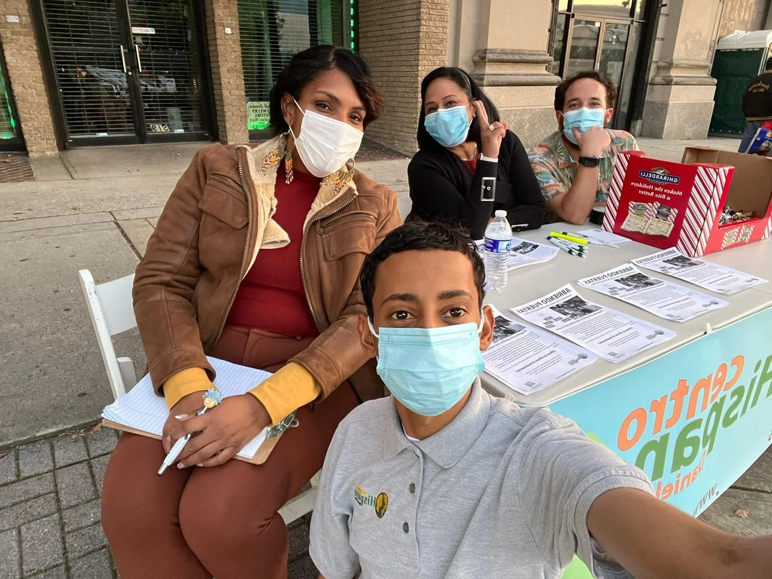 A group of four people wearing face masks are sitting at an outdoor table with pamphlets and a red box, possibly for community outreach or an informational event. The table has a banner that partially reads "Centro Hispano." They appear to be in a ci