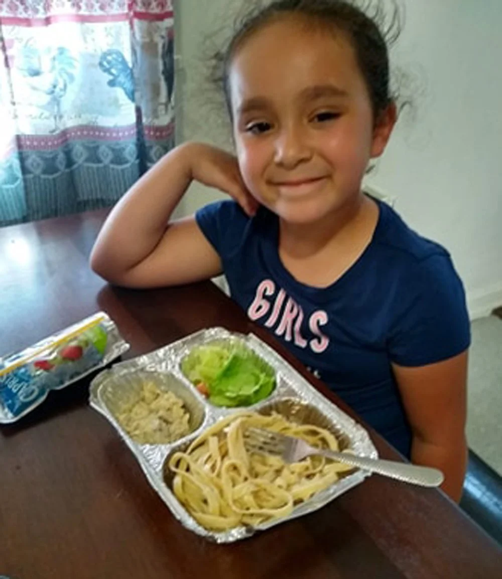 Child smiling at a table with a meal in a foil tray containing pasta, salad, and other items.