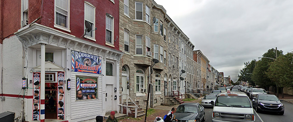 Street view of urban neighborhood with row houses and a barbershop. Several parked cars line the street under cloudy skies.