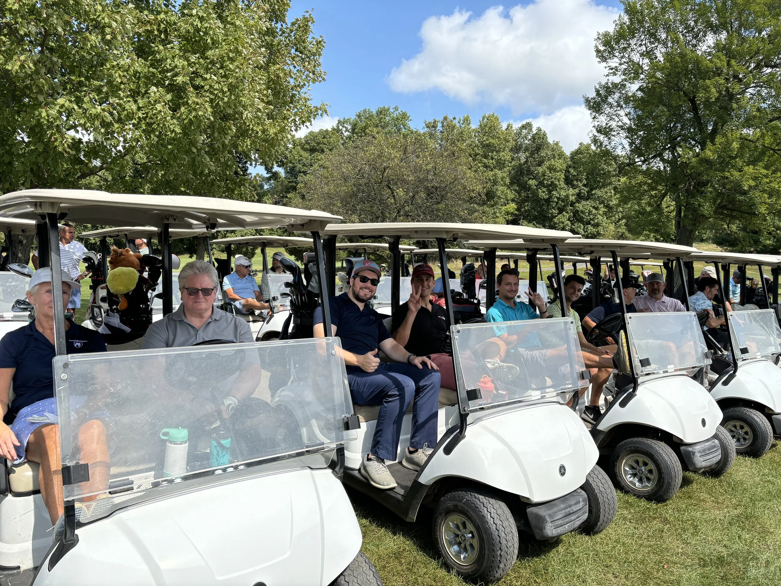 A group of people seated in a row of golf carts on a golf course, surrounded by trees and under a blue sky with clouds.