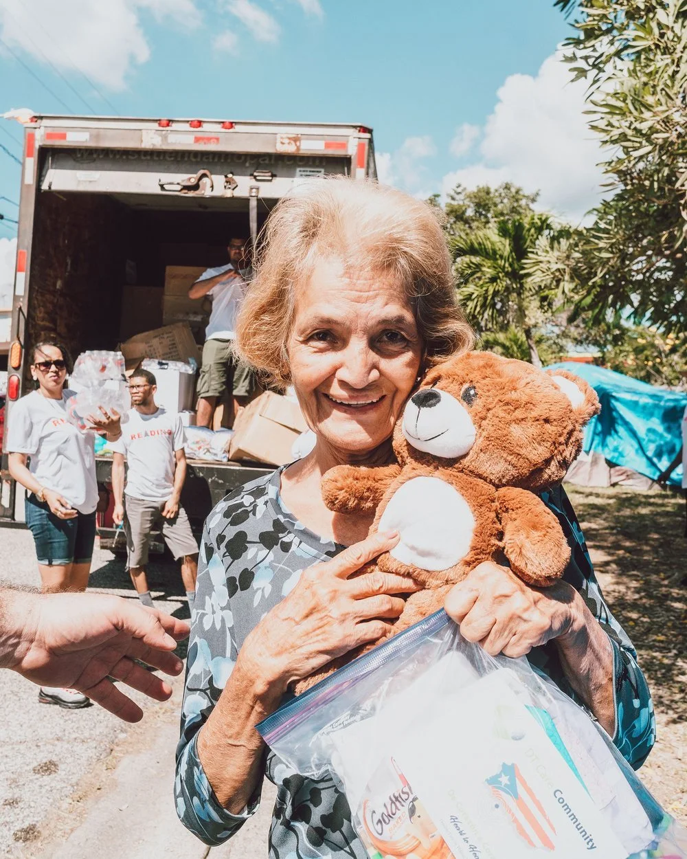 Elderly woman holding a teddy bear and a plastic bag containing relief supplies, with people unloading a truck of donations in the background.