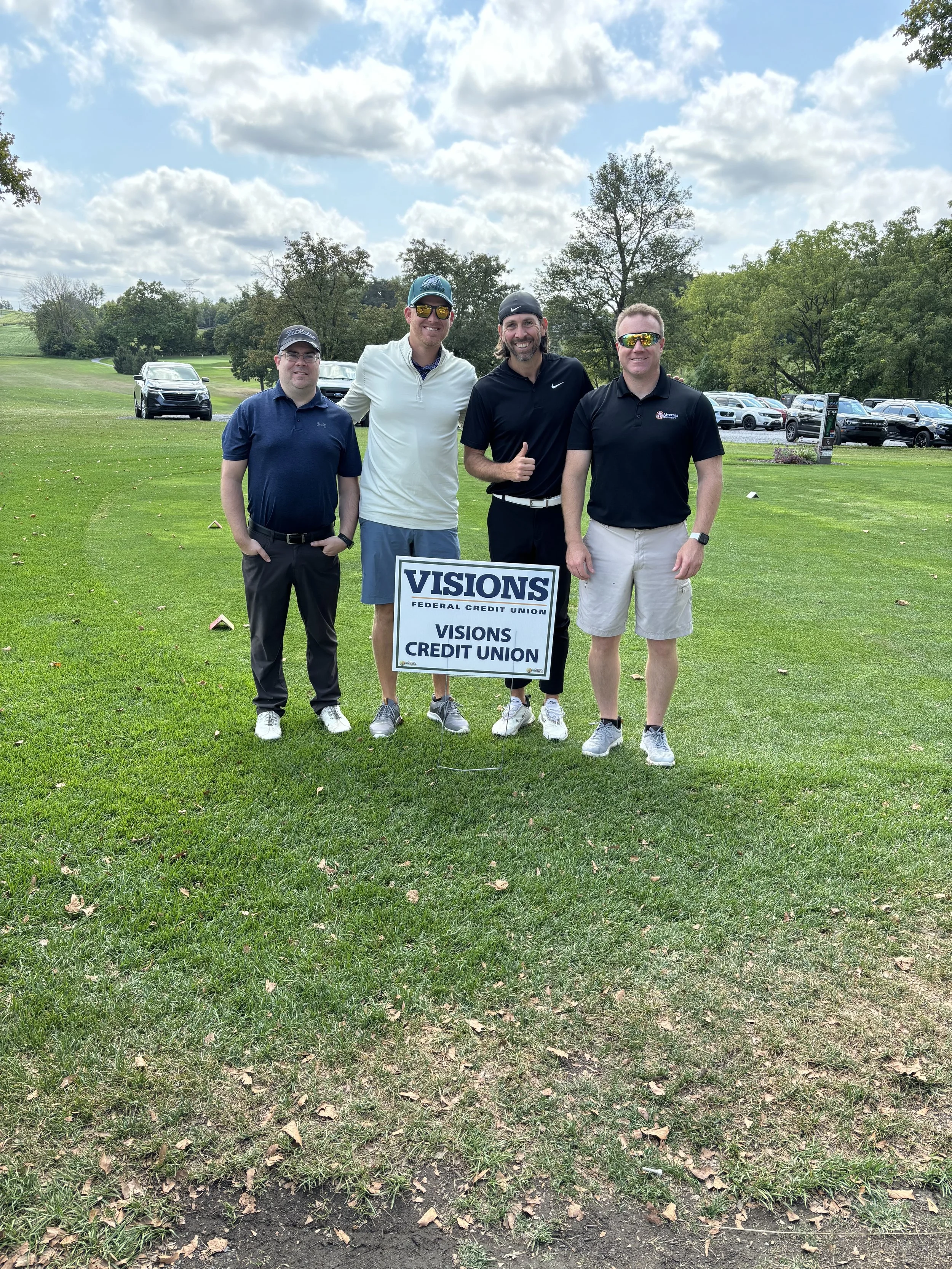 Four people standing on a grassy golf course holding a Visions Federal Credit Union sign with trees and parked cars in the background.