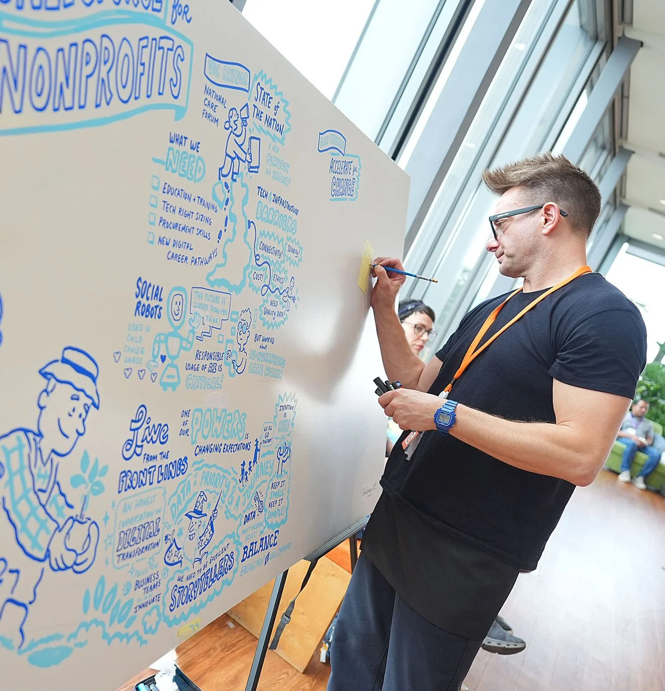 A man with glasses and a blue watch drawing on a large whiteboard with blue ink, with others in the background, in a well-lit indoor space with large windows.