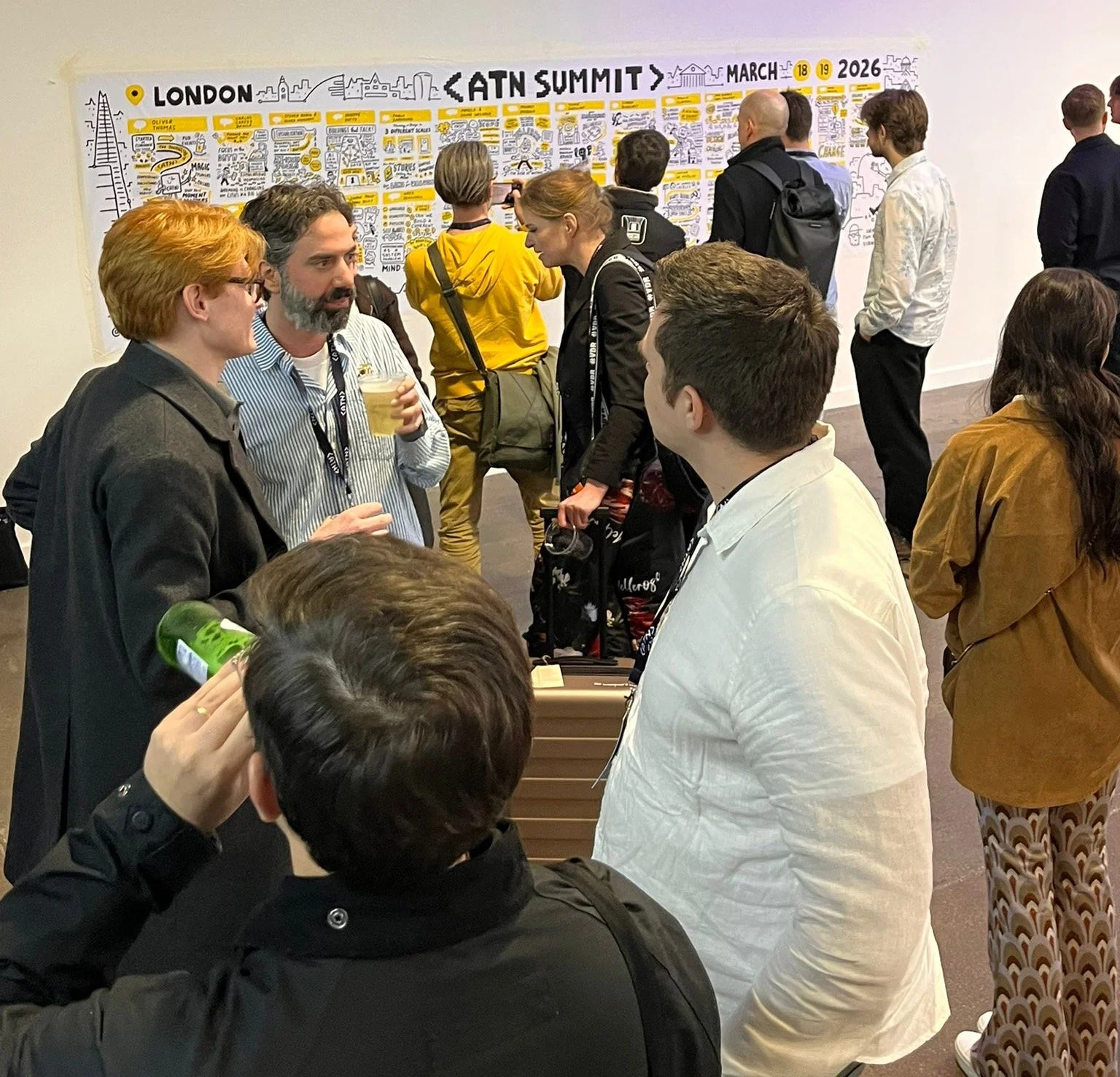 People attending an AI summit in London, March 18-19, 2026, are engaged in conversations near a large whiteboard with illustrations and text about the event.