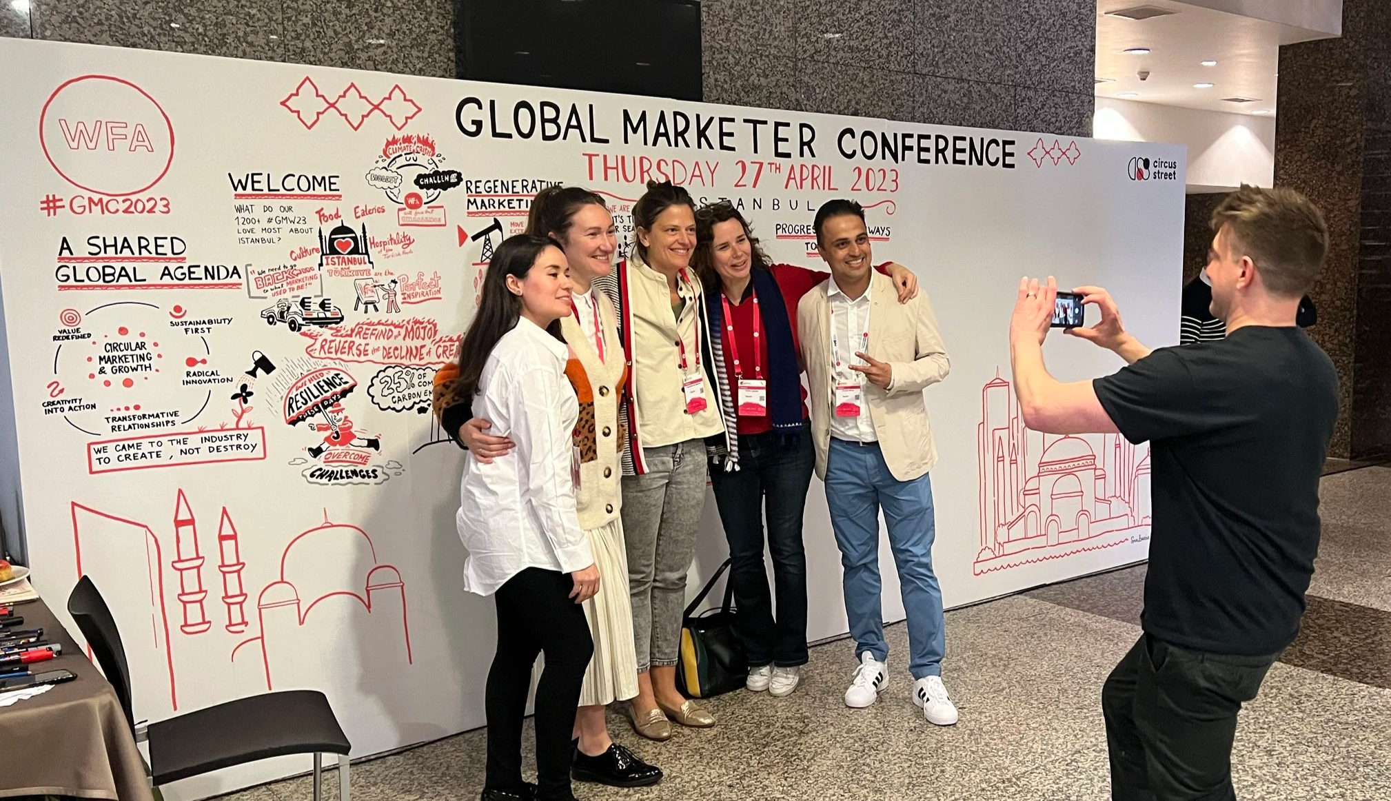 A group of five people posing for a photo in front of a conference backdrop with the title 'Global Marketer Conference' on April 27, 2023, while a person takes their picture with a smartphone.