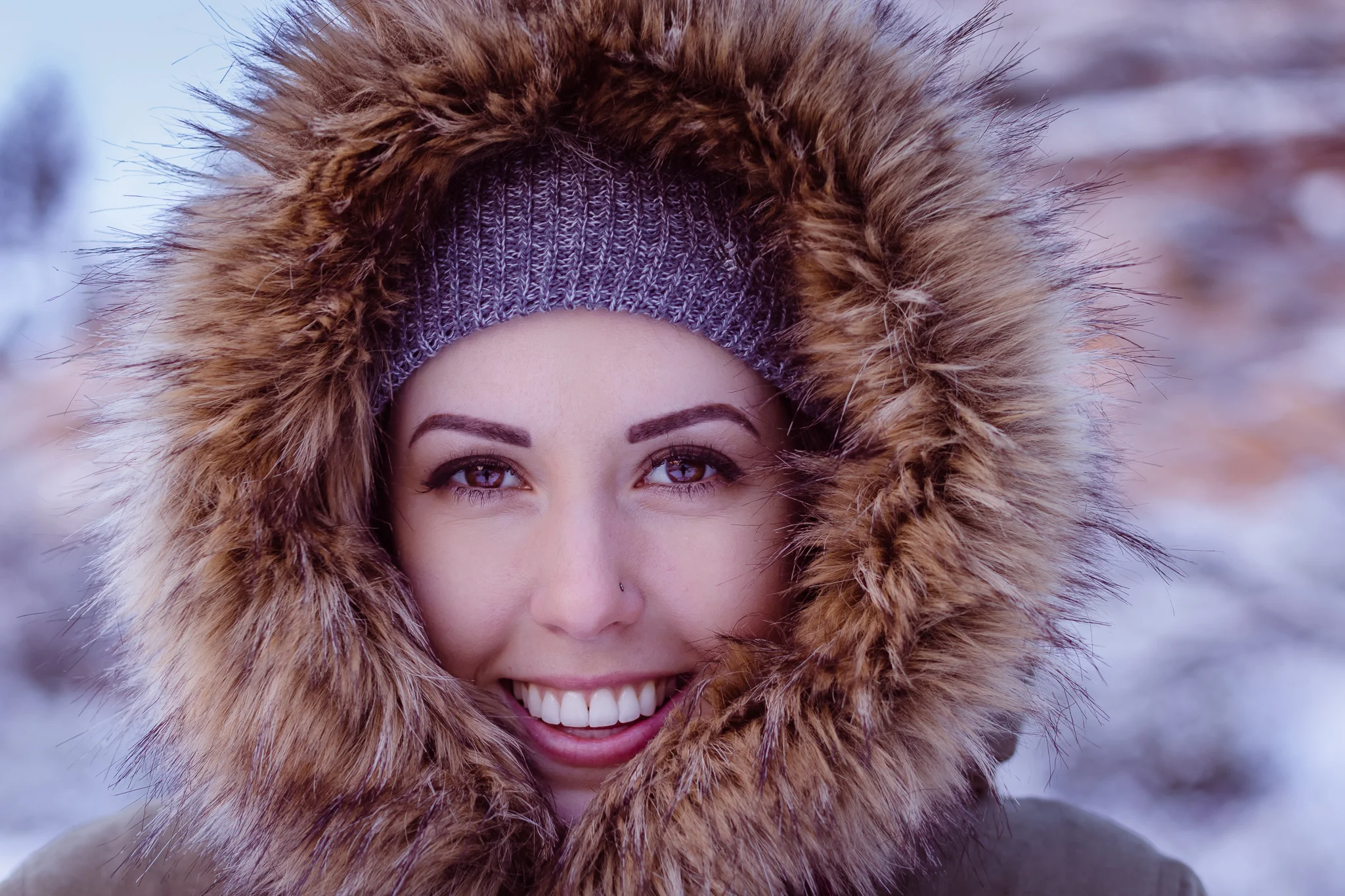 Close-up of a smiling woman wearing a fur-lined hood and a knit winter hat outdoors in a snowy landscape.